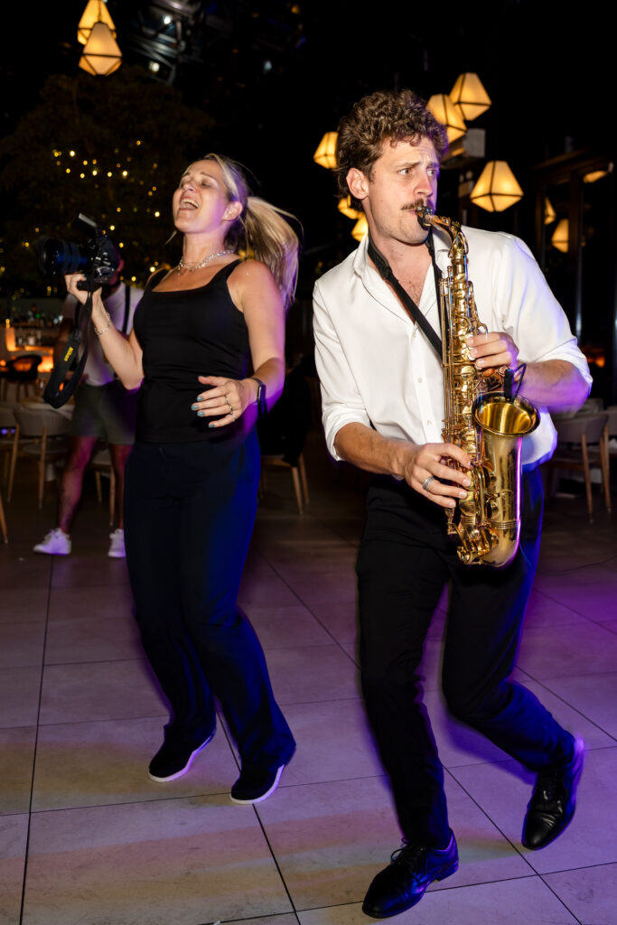 Wedding photographer dancing on the reception floor alongside a live saxophonist during a high-energy wedding celebration.