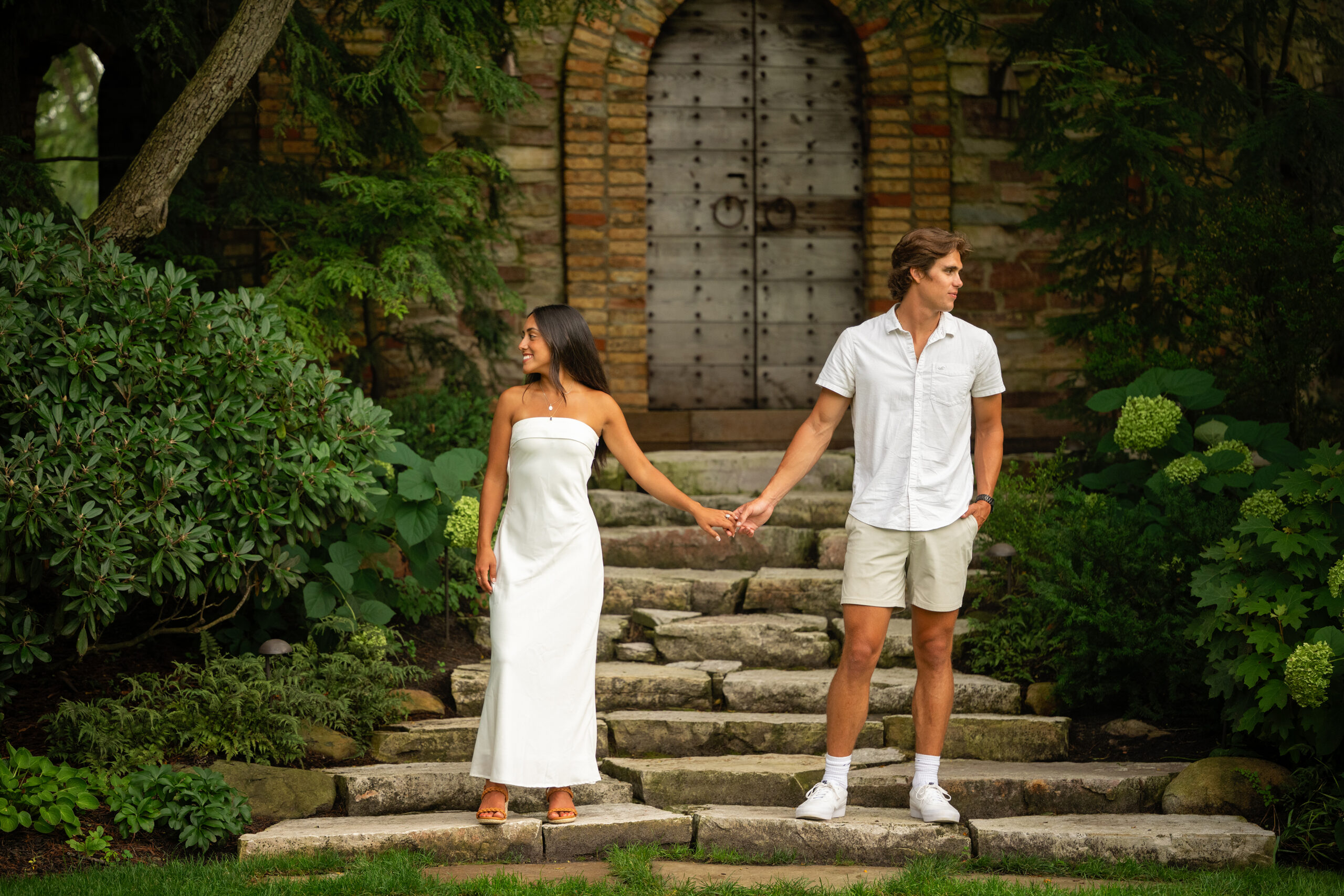 Young couple holding hands on stone steps in front of rustic doors at Hamstra Gardens engagement session